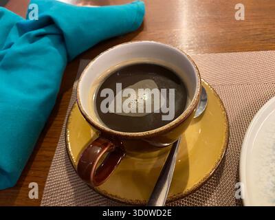 Black coffee with foam in a cup on a saucer with a spoon, with a turquoise napkin next to it Stock Photo