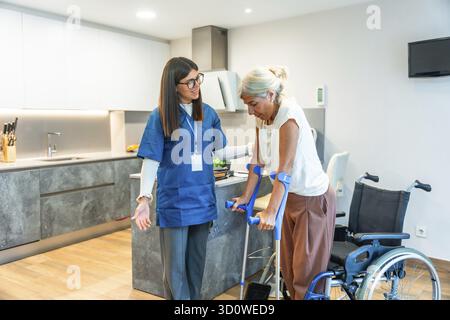 Home nurse assisting a senior woman using crutches to walk, providing support and physical therapy during her rehabilitation process in a modern home Stock Photo