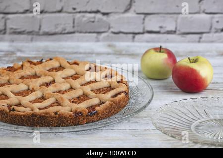 Apple cake on a glass plate with two apples next to it in front of a rustic brick wall Stock Photo