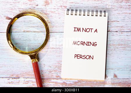 A notepad stating I am not a morning person sits beside a magnifying glass on a white wooden surface. Maybe tomorrow will bring a better day, just not Stock Photo