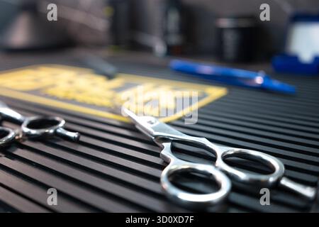 Barbershop tools arranged on a black rubber mat, featuring silver and black scissors, various combs, and a brush for professional hair styling. Stock Photo
