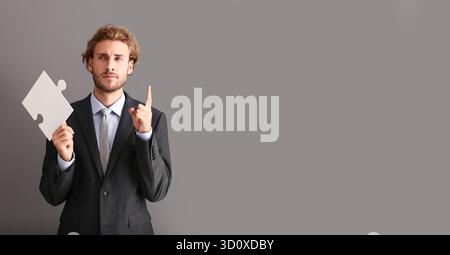 Thoughtful businessman with piece of jigsaw puzzle and raised index finger on grey background with space for text Stock Photo