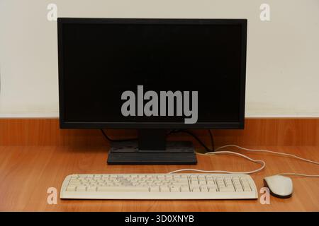 A simple workspace features a black monitor, white keyboard, and mouse on a wooden desk, creating a clean and organized look. Stock Photo