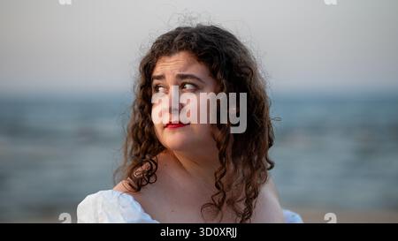 Young woman with curly hair gazes thoughtfully at the ocean during a serene beach sunset Stock Photo