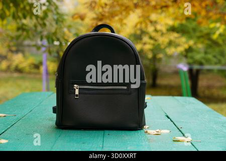 A stylish black backpack sits on a green wooden table with colorful autumn leaves in the background, showcasing a serene park setting. Stock Photo