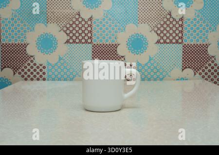 A plain white coffee mug stands alone on a polished surface, surrounded by a vibrant floral design. Stock Photo