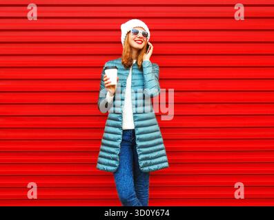 Fashion smiling woman talks on a smartphone holds coffee cup on red background || Model released Stock Photo