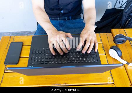 Person's hands typing on a laptop keyboard, working remotely from a wooden table with a smartphone and headphones Stock Photo