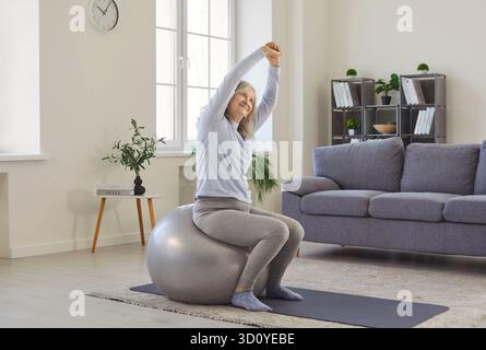 Old grey haired woman exercising with fitball, enjoying gymnastics at home, keeps arms above head Stock Photo