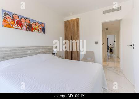 Simple white bedroom with wooden closet, decorative artwork, and view into hallway, showcasing a clean modern living space with minimal design. Stock Photo