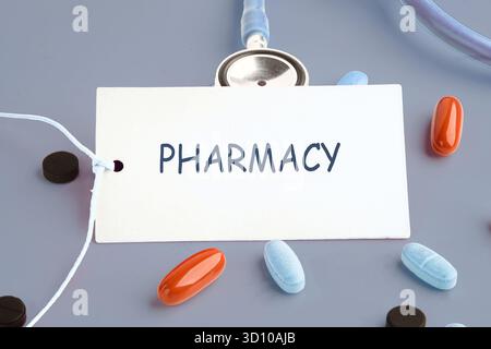 Pills of various colors are scattered around a white label that says Pharmacy. A stethoscope lies nearby on a grey surface. Medication and healthcare Stock Photo