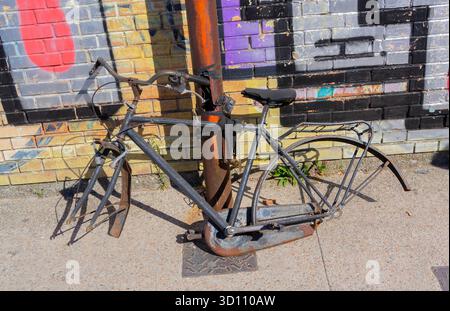 Old bicycle missing parts locked to rusty pole in front of colorful graffiti wall on sunny urban street. Stock Photo