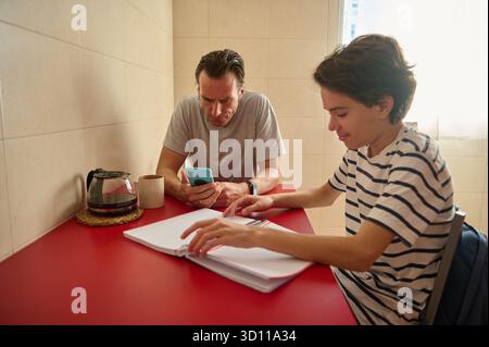 A father and son sit at a red table in a bright kitchen, one using a smartphone while the other writes in a notebook. The scene conveys learning, coll Stock Photo