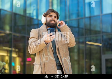 Happy Arabian Indian businessman putting on wireless headphones choosing, listening favorite energetic music in smartphone outdoors. Smiling man guy walking passes by urban city street. Lifestyles. Stock Photo