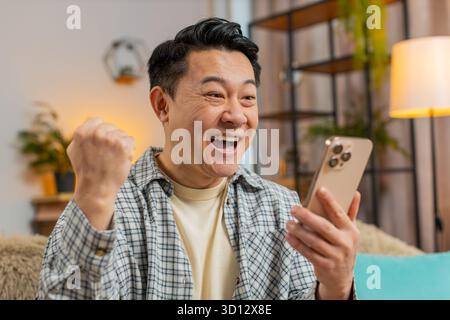 Asian middle-aged man sitting on sofa at home reading unexpected positive email news on smartphone. Surprised, celebrating online game victory. Happy, joyful man indoors enjoying successful moment Stock Photo