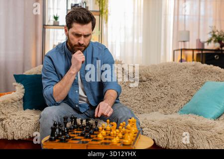 Young man sitting at home on sofa, playing chess alone, making a serious move with deep concentration. Caucasian guy strategizing, feeling thoughtful and focused, symbolizing dual personality indoors Stock Photo