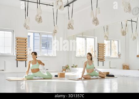 Two women are practicing stretching in a spacious and well-lit studio. They focus on different poses while enjoying the calm atmosphere created by nat Stock Photo