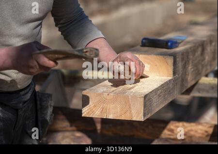 A carpenter skillfully shapes a wooden beam using a chisel and hammer in sunlight Stock Photo
