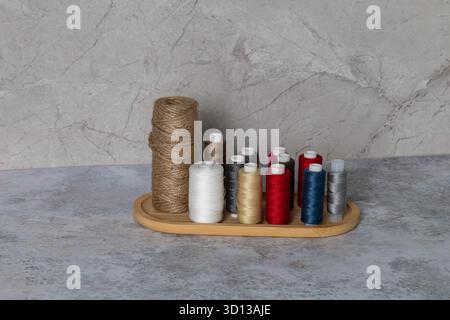 Spools of vibrant sewing thread displayed on a wooden tray against a textured gray backdrop, highlighting diverse colors and materials for crafting Stock Photo