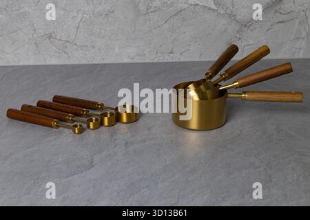 Brass measuring cups and spoons with wooden handles are elegantly arranged on a textured gray surface, highlighting essential kitchen tools Stock Photo