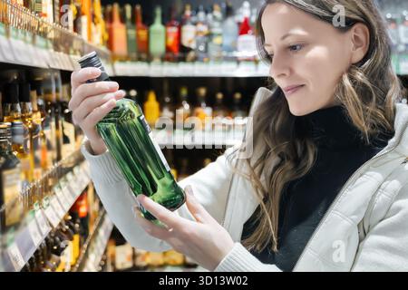 A young woman with long hair smiles while holding a green liquor bottle, deciding on a drink choice in the liquor department for an upcoming party. Stock Photo