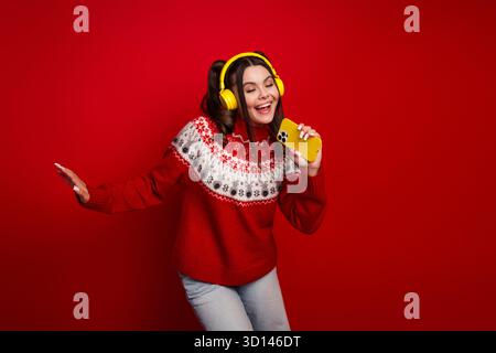 Young woman wearing a festive red sweater dances with joy while singing into a yellow smartphone with yellow headphones against a bold red background Stock Photo