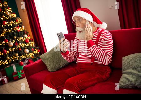 Santa Claus wearing a red striped outfit surprised while using a smartphone during Christmas season in a cozy home Stock Photo