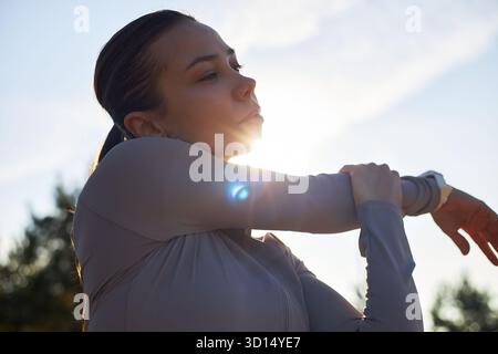 Young adult Caucasian woman stretching arm outdoors, looking away from camera, sunlight shining behind head, engaging in warm up exercise before physical activity, hair tied back Stock Photo