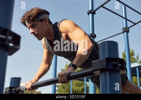 Caucasian young adult man exercising outdoors performing push ups on parallel bars, wearing fitness tracker focused on workout muscles engaged athletic training in outdoor gym Stock Photo