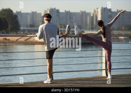 Caucasian young adult man and Caucasian young adult woman stretching legs and arms on waterfront promenade, exercising together outdoors with city buildings and river in background Stock Photo