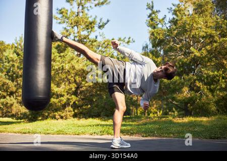 Caucasian young adult man performing high kick against punching bag outdoors, training martial arts in park, demonstrating athleticism and focus during workout session Stock Photo