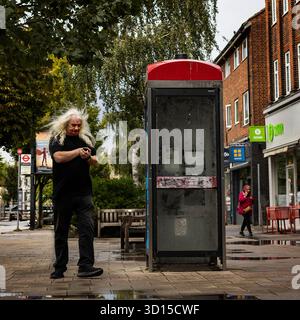 A long haired man walking down the street looking at his mobile phone as he walks past an old disused phone box Stock Photo