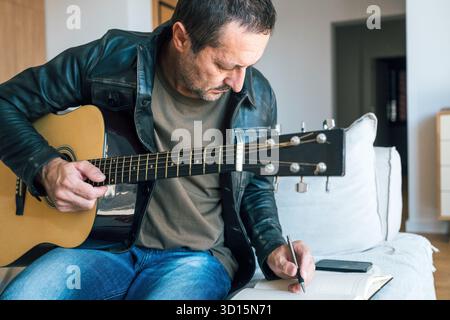 Mature man at home playing acoustic guitar and writing lyrics in notebook, concept of creativity, songwriting, and artistic inspiration in everyday li Stock Photo