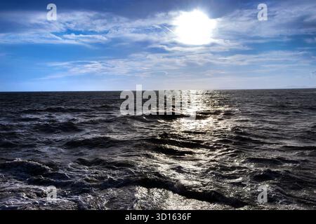 Sun reflecting over dark rippling ocean waves under a partly cloudy sky during late afternoon or early evening Stock Photo