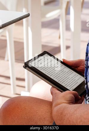 close up of woman reading a kindle e-book on a digital tablet while sitting outdoors with white chairs in the background C'an pastilla mallorca Stock Photo