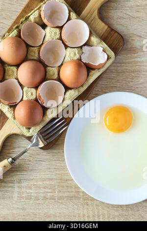 Overhead shot of whole and broken eggs on a wooden board next to a plate with a raw egg in a kitchen with natural light. Concept of food and homemade Stock Photo