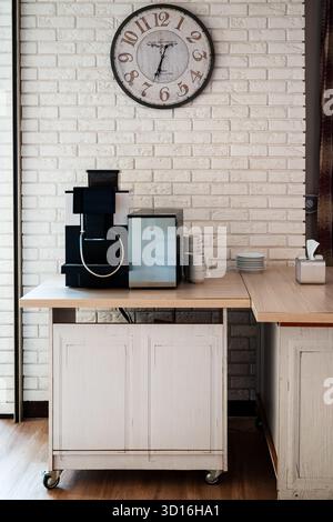 a coffee station at a hotel - coffee machine on a counter with cream white brick wall and decorative clock in the background Stock Photo