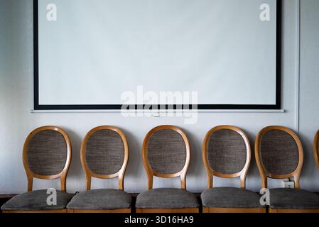 a row of stylized chairs under a blank white projector screen hanging on a wall - mock up photograph Stock Photo