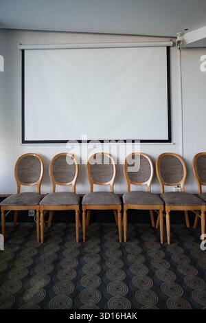 a row of stylized chairs under a blank white projector screen hanging on a wall - mock up photograph Stock Photo