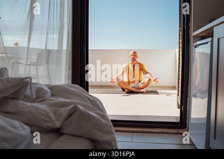 Middle-aged man doing morning yoga breathing exercises in Sukhasana easy pose on sunny summer terrace bedroom. Relaxed lifestyle concept with healthy Stock Photo