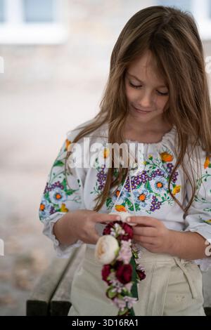 Vertical waist-up photo of a smiling Ukrainian girl in an embroidered blouse looking down at a floral wreath in her hands, concept of childhood joy an Stock Photo