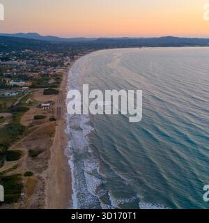Aerial panorama of a vast, curving sandy beach on Corfu island, Greece, during a soft pink sunset. Gentle waves create patterns in the turquoise water Stock Photo