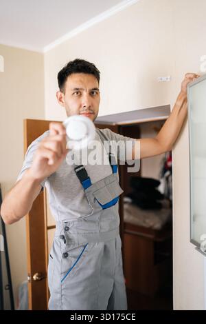 Vertical portrait of skilled electrician holding smoke detector while installing in residential building, prioritizing fire safety and ensuring Stock Photo