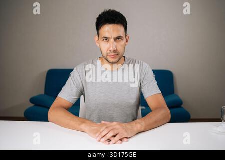 Indoor shot portrait of serious bearded man wearing casual gray t-shirt sitting at white table with clasped hands , conveying contemplation Stock Photo