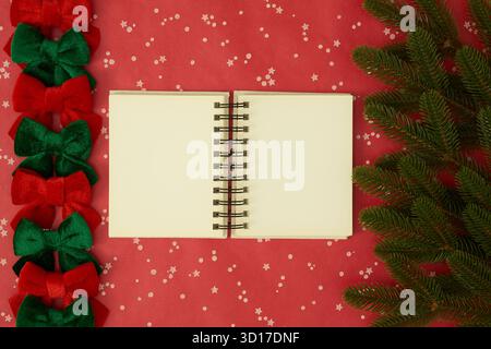 Open blank notebook in the center with Christmas tree branches and festive ribbons on a red background, holiday flat lay composition. Stock Photo