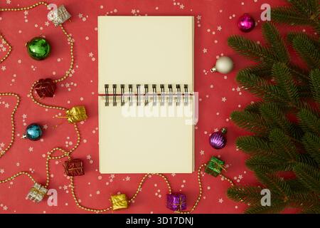 Open blank notebook in the center surrounded by Christmas tree branches, holiday decorations, and gifts on a red background Stock Photo