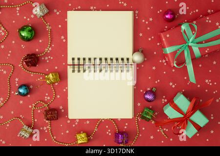 Open blank notebook in the center surrounded by Christmas tree branches, holiday decorations, and gifts on a red background Stock Photo
