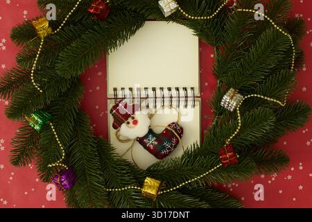 Open notebook framed by a Christmas wreath with red and green velvet bows, gift boxes, and gold beads on a red background Stock Photo