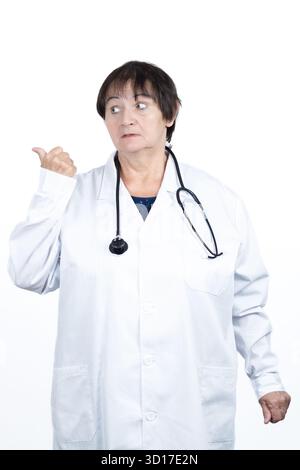 Female doctor in lab coat and stethoscope looks surprised and points her thumb toward background, studio shot on white with expression of concern Stock Photo
