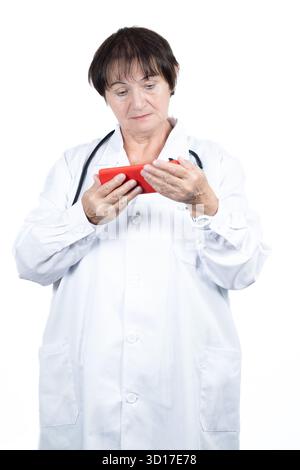 Senior female doctor wearing a white lab coat and stethoscope, focused on using a red smartphone, representing telemedicine, digital healthcare Stock Photo